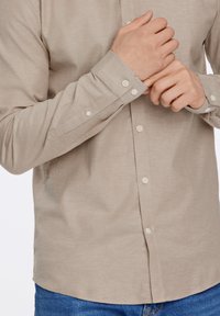 Camicia beige a maniche lunghe con design a bottoni, caratterizzata da una sottile texture e bottoni bianchi, abbinata a jeans blu.