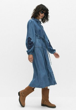 Femme marchant dans une robe en denim bleue mi-longue avec des manches bouffantes et une broderie foncée, portant des bottes marron mi-mollet et des lunettes de soleil noires.