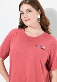 Camiseta de color rosa con mangas cortas y escote redondo, que presenta "OPTimIST by PURE" bordado en multicolores en el pecho.
