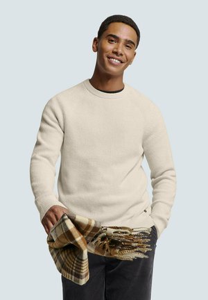 Crème-kleurige gebreide trui met een ronde hals. Model houdt een bruin en beige geruit sjaal met franjes vast. Achtergrond is lichtblauw.