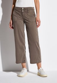 Pantalones sueltos hasta el tobillo, de color gris-marrón, con dos bolsillos laterales, cuatro botones y detalles de costura visibles. Llevados con zapatillas blancas.