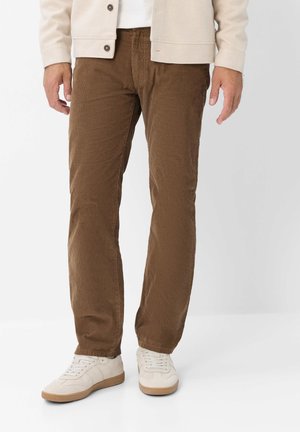 Uomo che indossa pantaloni di velluto a coste marroni, giacca beige e sneakers bianche, in piedi davanti a uno sfondo semplice.
