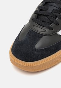 Zwarte sneaker met bovenwerk van suede en leer, textuur gumzool, ronde neus en platte veters. Heeft een strakke vormgeving met subtiele accenten.