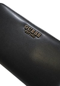 Czarny skórzany portfel z wytłoczonym logo "GUESS EST 1981" w złotym kolorze. Gładka faktura, prostokątny kształt i elegancki design.