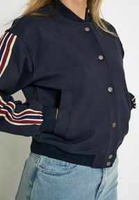 Navy bomber jacket with snap buttons, striped accents in red and cream, and ribbed cuffs. Features side pockets and a smooth texture.