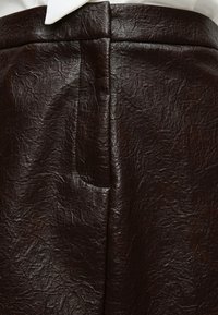 Gros plan d'une jupe en cuir texturé marron foncé avec un détail de couture visible et une partie d'un col de chemise blanche au-dessus de la ceinture.