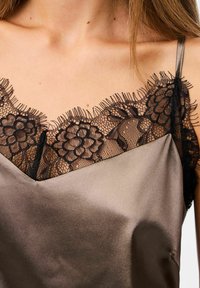 Camisole in satin di una ricca tonalità marrone, con delicati bordi in pizzo nero lungo il collo, decorato con intricate fantasie floreali e spalline regolabili.