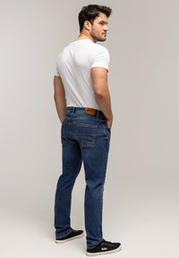 Mężczyzna w białej koszulce i niebieskich dżinsach stoi z rękami w kieszeniach, patrząc przez ramię, na jednolitym jasnym tle.