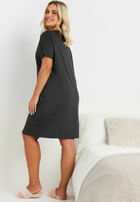 Abito t-shirt grigio scuro con maniche corte, di lunghezza al ginocchio e una vestibilità comoda. Indossato con pantofole morbide rosa chiaro, ambientato su uno sfondo di letto.