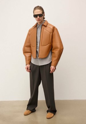 Pruunist nahast lühendatud jakk, millel on polsterdatud õlad, hall ribadest särk, tumedad laiad püksid ja koorikujulised jalanõud, neutraalse taustaga.