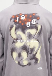 Πίσω μέρος γκρι φούτερ με κουκούλα με αφηρημένο μοτίβο σε πορτοκαλί, μπεζ και μαύρο, λευκό κείμενο «NEVER STOP EXPLORING» και το λογότυπο The North Face.