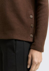 Pull en tricot marron avec un tissu texturé et trois boutons décoratifs sur le côté. Pantalon noir partiellement visible en dessous.