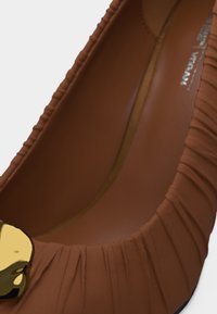 Chaussure plate marron froncée avec une décoration en forme de cœur doré brillant près de l'ouverture des orteils.