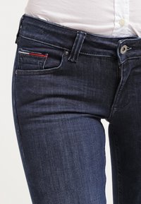 Jeans de mezclilla de color azul oscuro con un corte ajustado, un leve desgastado y un pequeño acento rojo en el bolsillo delantero. Diseño clásico de cinco bolsillos.