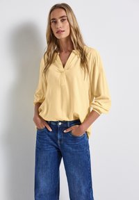 Blusa gialla con scollo a V profondo e maniche ampie, abbinata a jeans in denim blu con tasche frontali e chiusura con bottone.