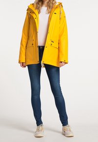 Veste imperméable jaune vif avec capuche, fermeture éclair, deux poches avant et détails à cordon, associée à un jean bleu foncé et des baskets.