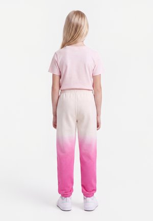 Enfant aux longs cheveux blonds, debout de dos, portant un t-shirt rose clair et un pantalon de jogging ombré rose vers blanc avec des baskets blanches.