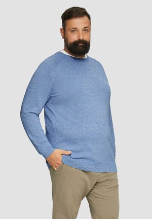 Niebieski sweter dzianinowy z długimi rękawami, okrągłym dekoltem i fakturowanym wzorem. Połączony z jasnymi spodniami. Model stoi z rękami w kieszeniach.