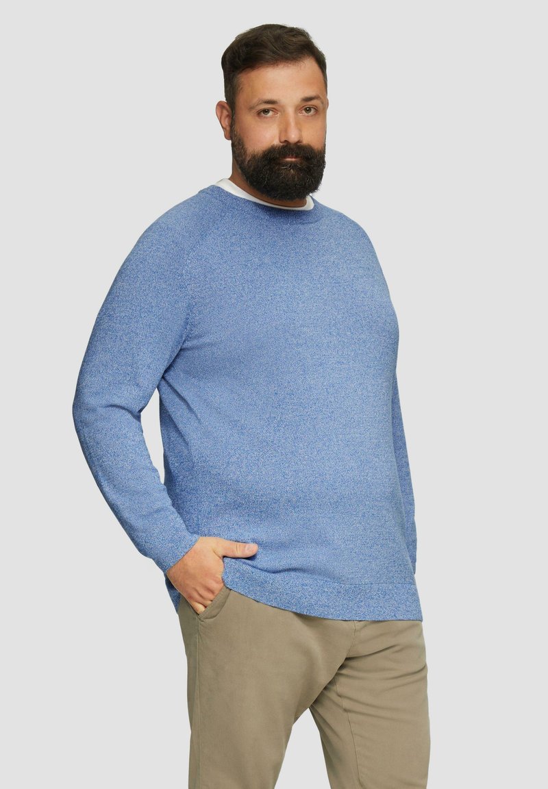 Blauer gestrickter Pullover mit langen Ärmeln, Rundhalsausschnitt und strukturiertem Muster. Kombiniert mit hellen Hosen. Modell steht mit den Händen in den Taschen.
