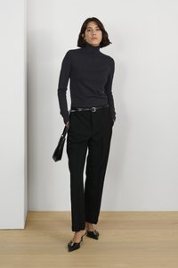 Un pull noir à col roulé, un pantalon noir ajusté et des escarpins noirs à bouts pointus. Assorti à un petit sac noir, sur un sol en bois.