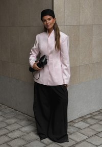 Sieviete, ģērbta gaiši rozā žaketē, platās melnās biksēs, melnā beretē un turot melno somiņu, stāv pie flīžu sienas stūra.