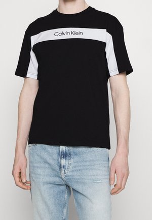 Mężczyzna w czarno-białej koszulce Calvin Klein z krótkim rękawem, zestawionej z jasnoniebieskimi dżinsami na jednolitym tle.
