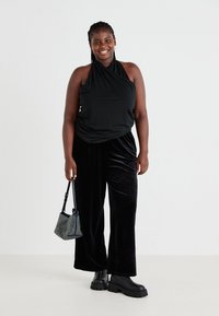 Top halter negro y pantalones de pata de elefante de terciopelo negro. La modelo sostiene un pequeño bolso con detalles metálicos. Botas negras de plataforma gruesa completan el conjunto.