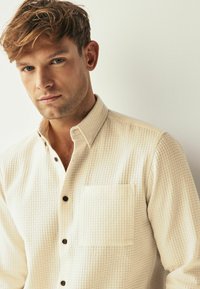 Krèmfarget skjorte med knappelukking, teksturert mønster, krage og frontlomme. Brune knapper står i kontrast til det lyse stoffet.