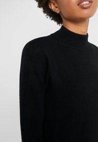 Svart ribbad polotröja med en figursydd design. Har långa ärmar och en slät textur som framhäver en enkel, modern estetik.