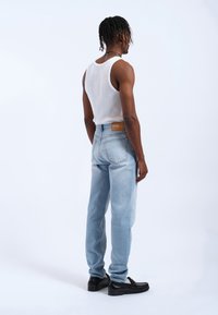 Jeans de mezclilla azul claro con un corte relajado, bolsillos traseros y una etiqueta marrón en la cinturilla, combinados con una camiseta sin mangas blanca y zapatos negros.