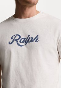 White cotton t-shirt with a round neckline, featuring a distressed blue "Ralph" logo printed on the front. Smooth texture, casual design.