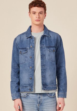Leren spijkerjack in lichtblauw met een klassiek ontwerp, knoopsluiting en twee borstzakken. Gedragen over een lichtgrijze T-shirt met ronde hals.