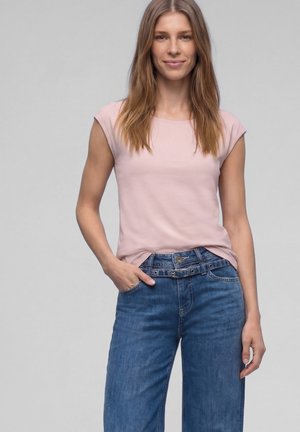 Femme aux cheveux raides châtain clair portant un t-shirt rose clair à manches courtes et un jean bleu avec une ceinture à la taille, debout avec une main dans la poche.