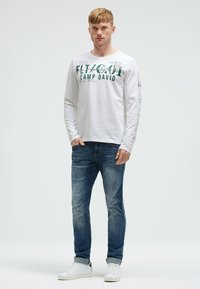 White long-sleeve shirt with green lettering, paired with blue denim jeans and white sneakers. The shirt features printed designs on sleeves.
