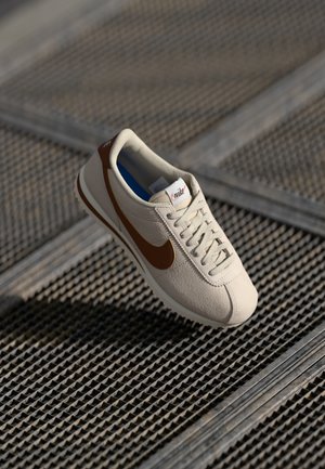 Beige Nike-sneakers met een bruine swoosh en veters geplaatst op een getextureerd metalen oppervlak.