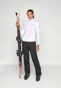 Balts garām piedurknēm slēpošanas krekls ar rozā akcentiem, kopā ar melnām slēpošanas biksēm un melniem slēpošanas zābakiem; rokās tur slēpes ar rozā un melnu dizainu.