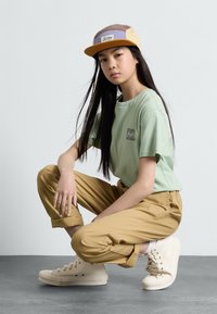 T-shirt en coton vert clair avec un petit graphique, pantalon cargo kaki coupé, et baskets crème montantes, le tout sur fond uni.