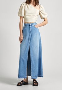 Kvinna iklädd en beige randig puffärmad croptopp, högmidjade ljusblå denimmaxikjol med slits fram och svarta sandaler med spänne.