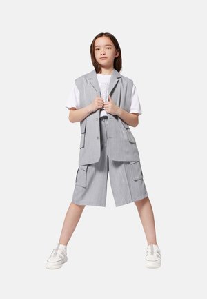 Ensemble en gris à rayures avec gilet et short, doté de poches, associé à un t-shirt blanc. La modèle se tient avec confiance dans des baskets blanches.