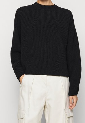 Person iført en sort striktrøje med lange ærmer og hvide cargobukser, den ene hånd i lommen, mod en ensfarvet baggrund.