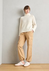 Maglione a collo alto a coste crema, pantaloni cargo beige con tasche laterali e sneakers bianche con dettagli rosa, il tutto su uno sfondo neutro.