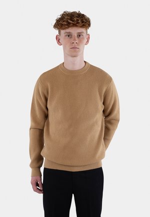 Beige ribbestrikket genser med rund hals og lange ermer, med en løs passform. Brukt med mørke bukser mot en lys bakgrunn.