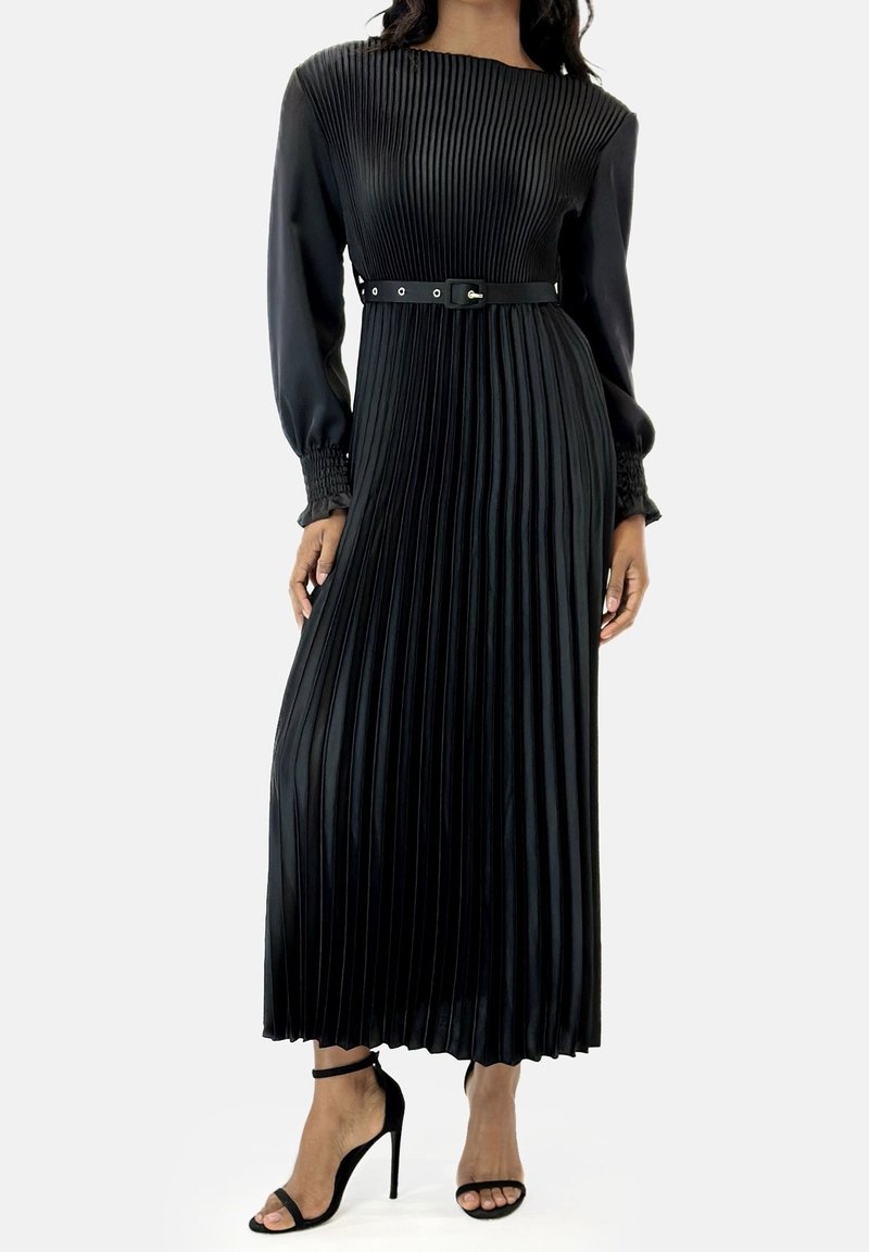 Femme portant une longue robe noire plissée avec une ceinture à la taille et de longues manches bouffantes, associée à des sandales noires à talons hauts.