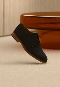 Μαύρο δερμάτινο παπούτσι με κορδόνια, με λείο ύφασμα στο πάνω μέρος, στρογγυλεμένη μύτη και ελαφριά καφέ ξύλινη σόλα. Διαθέτει διακριτική ραφή και λεπτομέρεια με λογότυπο.