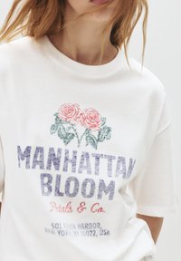 Bílý bavlněný tričko s květinovou grafikou v růžové a zelené barvě, text "MANHATTAN BLOOM" v modré, kulatý výstřih a uvolněný střih.