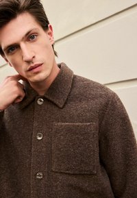 Veste en laine brune avec un col, des boutons sur le devant et une poche poitrine. La texture est douce et légèrement duveteuse.