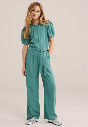 Ragazza con capelli lunghi biondi e occhiali rotondi, che indossa una tuta verde acqua con bottoni, cintura, maniche a sbuffo e scarpe argento, in piedi davanti a uno sfondo semplice.