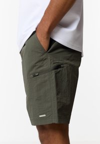 Άνδρας που φοράει χακί βερμούδα με πλαϊνή τσέπη με φερμουάρ και λευκό πουκάμισο, με το χέρι του μέσα στην μπροστινή τσέπη.