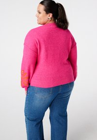 Fuchsia gebreide trui met hoge hals, met een geborduurd zonnestraalontwerp op de mouwen. Gecombineerd met blauwe jeans met rechte pijpen.