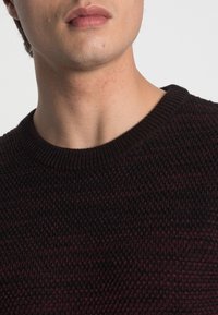 Stickad tröja med rundhals. Har ett texturerat mönster i djup burgund och svart. Mjukt, varmt material med ribbade ärmslut.